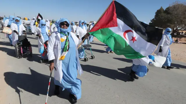 Une femme âgée portant un foulard bleu et blanc porte un drapeau du Sahara occidental devant d'autres manifestants